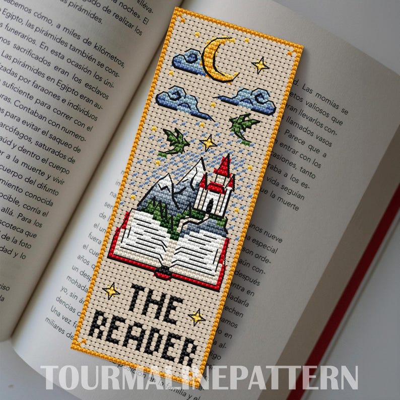 Fantasy cross-stitch bookmark: open book grows into mountains and a red-roof castle under a crescent moon, clouds, tiny stars, and two small dragons; “THE READER” text at the bottom, golden border. TourmalinePattern on Etsy