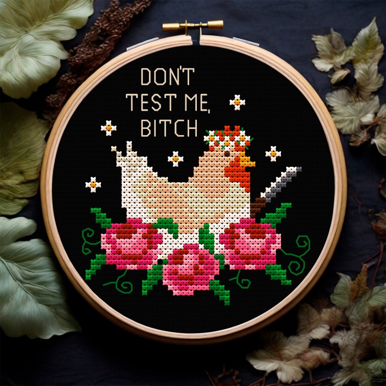 Puede incluir: Bordado de punto de cruz en un aro de madera con un pollo con corona de flores y un cuchillo, rodeado de rosas rosas y hojas verdes. El texto "DON'T TEST ME, BITCH" est&aacute; bordado sobre un fondo negro.