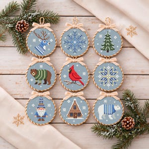 Peut inclure: Neuf ornements circulaires en point de croix sur une surface en bois clair. Chaque pièce présente un fond bleu clair avec des motifs hivernaux : cerf, flocon, sapin, ours, cardinal, étoile géométrique, gnome, chalet et ours polaire, avec des nœuds beiges.