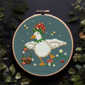 May include: Cross-stitch embroidery of a white goose wearing a red mushroom hat and holding a basket of red berries. The goose is surrounded by green leaves, white flowers, and red ladybugs on a blue background.