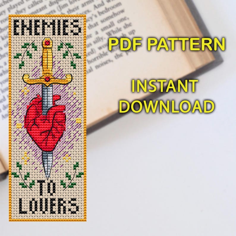 Cross stitch bookmark featuring a red anatomical heart pierced by a golden sword, surrounded by decorative leaves and sparkles. The text 'Enemies to Lovers' is stitched in bold black lettering above and below the central motif.