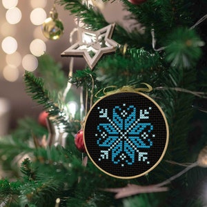 May include: A black cross-stitch Christmas tree ornament featuring a blue and white snowflake design. The round ornament is framed in a wooden hoop and suspended by a gold ribbon. The tree is decorated with other festive ornaments.
