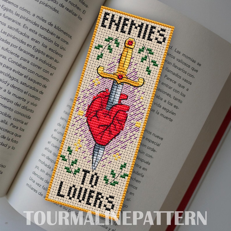 Cross stitch bookmark featuring a red anatomical heart pierced by a golden sword, surrounded by decorative leaves and sparkles. The text 'Enemies to Lovers' is stitched in bold black lettering above and below the central motif.