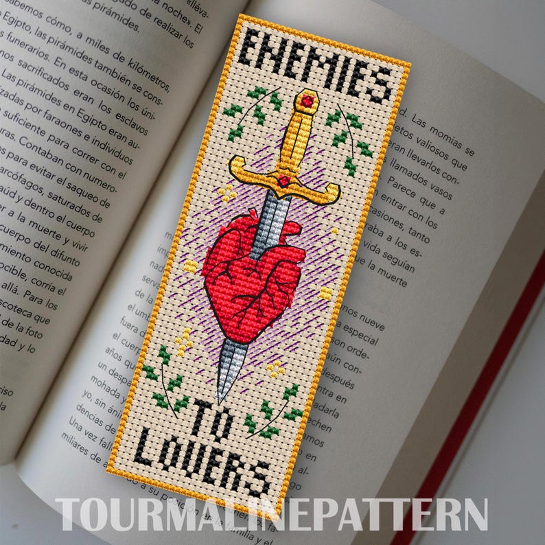 Bookmark on beige Aida: gold sword motif over a stylized red heart, purple hatch marks, green sprigs, and the phrase “ENEMIES TO LOVERS.” TourmalinePattern on Etsy