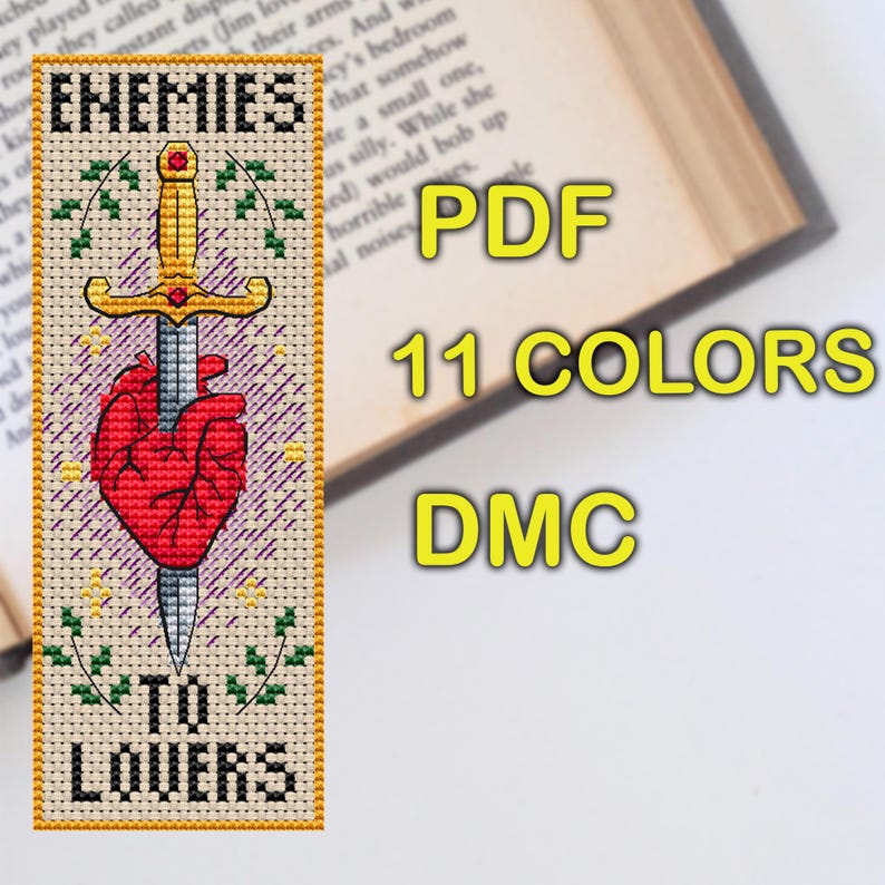 Cross stitch bookmark featuring a red anatomical heart pierced by a golden sword, surrounded by decorative leaves and sparkles. The text 'Enemies to Lovers' is stitched in bold black lettering above and below the central motif.