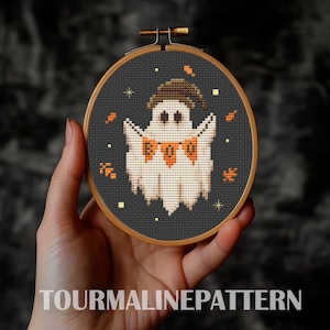 May include: A cross-stitch embroidery hoop with a black background and a white ghost with a black hat holding a banner that says "BOO". The ghost is surrounded by orange and yellow leaves and stars.