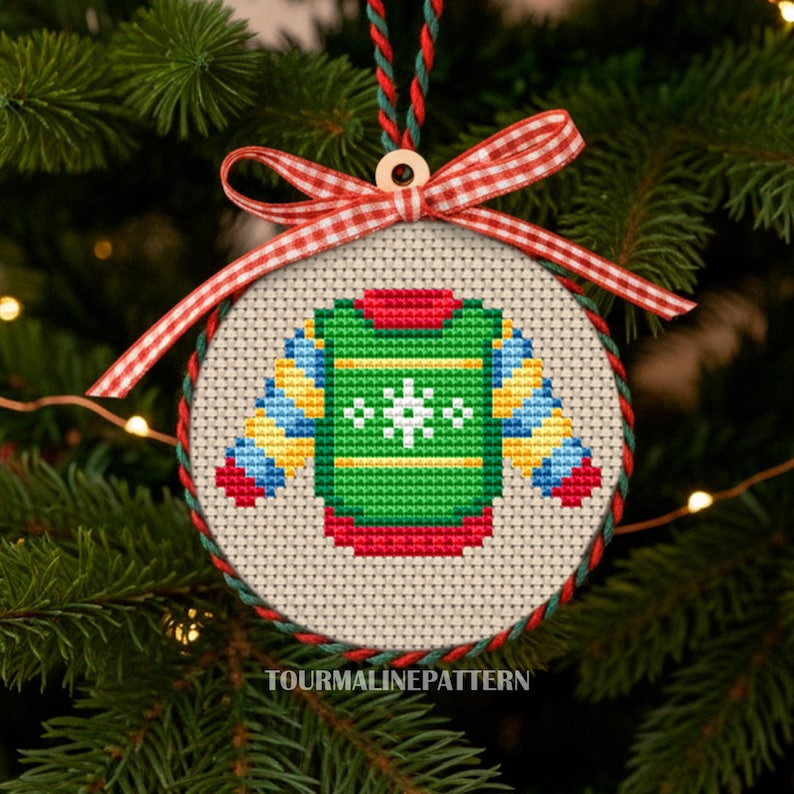 Christmas ornament cross stitch of a green sweater with snowflake in round hoop with red gingham ribbon on tree, TourmalinePattern on Etsy.