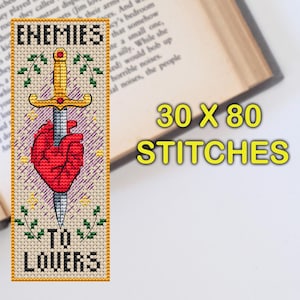 Cross stitch bookmark featuring a red anatomical heart pierced by a golden sword, surrounded by decorative leaves and sparkles. The text 'Enemies to Lovers' is stitched in bold black lettering above and below the central motif.
