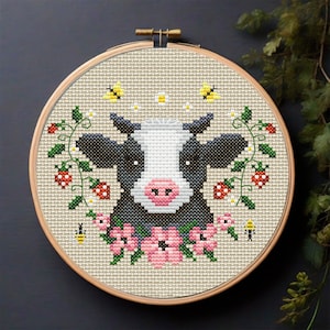 Puede incluir: Aro de bordado de punto de cruz con una vaca negra y blanca con la nariz rosa rodeada de flores rosas, hojas verdes y abejas amarillas.