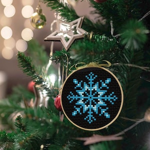 May include: A blue snowflake cross-stitch ornament hanging on a Christmas tree. The snowflake is embroidered on black fabric and framed with a wooden hoop.