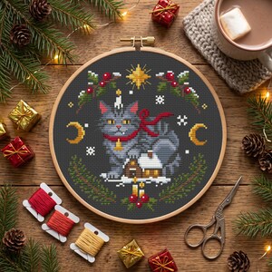 May include: A circular wooden embroidery hoop containing a cross-stitch pattern of a grey cat with a red ribbon, a small house, a yellow star, and two crescent moons on a dark grey background, surrounded by festive holiday decorations.