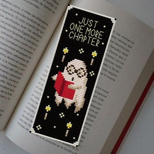 Puede incluir: Un marcapáginas de punto de cruz en blanco y negro con un fantasma leyendo un libro rojo con el texto "Just One More Chapter" en blanco. El fantasma está rodeado de pequeñas estrellas amarillas.