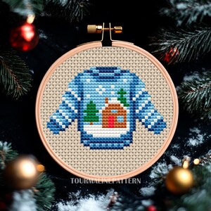 Christmas embroidery hoop with a blue sweater showing a winter landscape with house and trees, hanging on snowy tree, TourmalinePattern on Etsy.