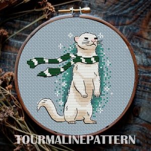 May include: A cross-stitch pattern of a white ferret wearing a green and white scarf. The ferret is standing on its hind legs with its tail curled behind it. The background is a light blue with green and white snowflakes.