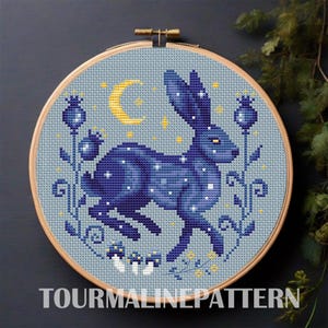 Puede incluir: Bordado de punto de cruz en un aro de madera con un conejo azul oscuro con detalles blancos en forma de estrella, una luna creciente amarilla y detalles florales. El texto "TOURMALINEPATTERN" está en la parte inferior. El diseño tiene un fondo azul claro.