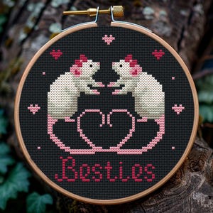 Könnte beinhalten: Stickrahmen mit Kreuzstich-Stickerei auf schwarzem Hintergrund, auf dem zwei weiße Ratten mit rosa Ohren und Nasen abgebildet sind, die sich gegenüberstehen. Die Ratten sind von rosa Herzen umgeben und das Wort "Bestiees" ist darunter in rosa gestickt.