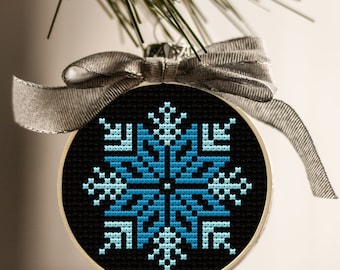 May include: A round Christmas ornament with a black background and a blue snowflake design. The ornament is framed in a wooden hoop and has a silver ribbon for hanging. The ornament is hanging from a pine branch.