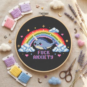 Può includere: Un telaio da ricamo a punto croce con un narvalo sotto un arcobaleno e la scritta "FUCK ANXIETY". L'immagine include rocchetti di filo, forme in feltro e lavanda. Il telaio ha un diametro di circa 20 cm.