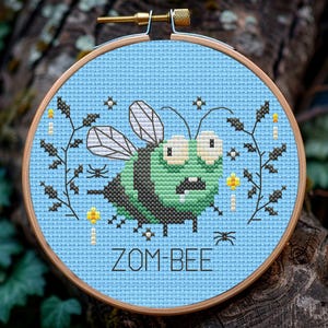 May include: A cross-stitch embroidery hoop with a blue background featuring a green zombie bee with black stripes and the text "ZOM-BEE".
