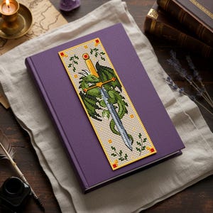 Cross-stitch bookmark with a green dragon coiled around a silver longsword; gold hilt with red gems, ivy sprigs, small gold stitches, and a yellow border on beige fabric—vertical fantasy design for readers. TourmalinePattern on Etsy
