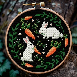 May include: A black embroidery hoop with a white background featuring two white rabbits and orange carrots. The rabbits are surrounded by green leaves and white flowers.