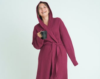 May include: A woman wearing a burgundy waffle-weave robe with a hood. She is holding a black mug in her right hand and smiling.