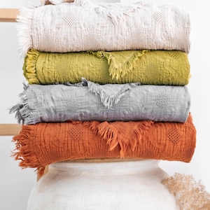May include: A stack of four rectangular blankets with fringed edges, each featuring a textured diamond pattern. The blankets are colored in light beige, olive green, light gray, and burnt orange, and are folded neatly on top of each other.