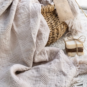 Peut inclure: Une couverture en coton beige douce avec un motif tissé et des franges sur les bords. La couverture est posée sur un panier en osier avec une étiquette indiquant "100% coton".