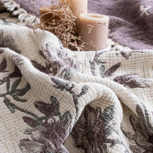 May include: A cream-colored quilted blanket featuring a dark purple and muted green floral pattern. The fabric has a textured, puckered surface. In the background, two beige pillar candles and dried foliage rest on a folded purple throw.