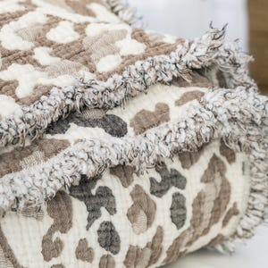 May include: A folded blanket with a cream-colored background and a brown and gray animal print pattern. The blanket has a fringed edge. The blanket is made of a soft, textured material.