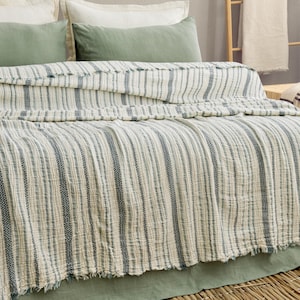 Pu&ograve; includere: Un letto con una coperta a righe, bianco sporco e blu. La coperta ha un bordo sfrangiato ed &egrave; drappeggiata su un letto con cuscini verdi e un copriletto verde. Il letto &egrave; in una stanza con una scala di legno e un tavolo di legno.