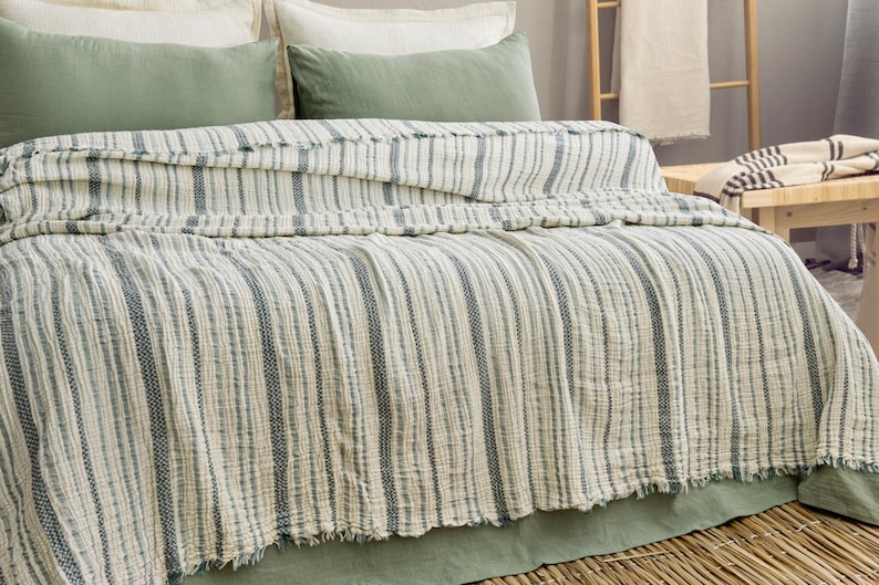 Pu&ograve; includere: Un letto con una coperta a righe, bianco sporco e blu. La coperta ha un bordo sfrangiato ed &egrave; drappeggiata su un letto con cuscini verdi e un copriletto verde. Il letto &egrave; in una stanza con una scala di legno e un tavolo di legno.