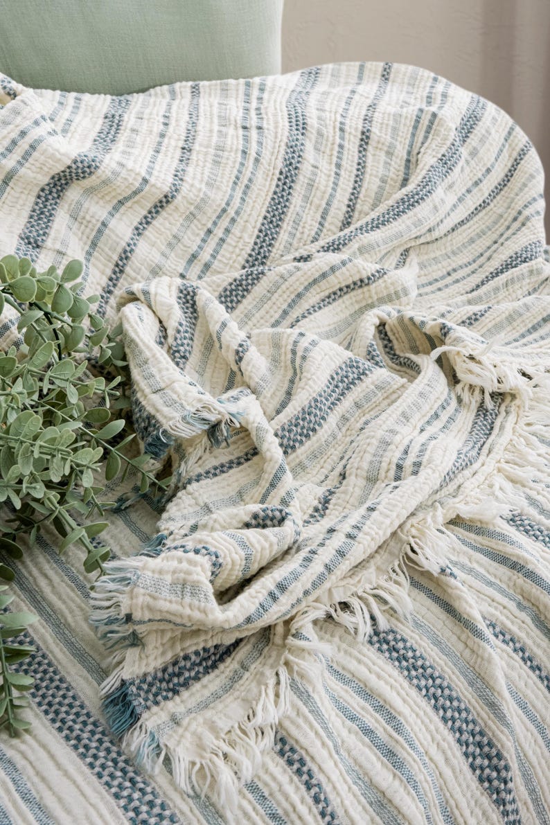 Pu&ograve; includere: Una coperta color crema con strisce blu e frange. La coperta &egrave; drappeggiata su una sedia, con un aspetto morbido e strutturato. Il design presenta strisce blu alternate, a tinta unita e fantasia. Una pianta verde &egrave; nell'angolo.