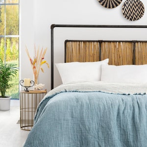 Peut inclure: Une couverture en coton bleu clair avec un tissage texturé et des bords effilochés. La couverture est drapée sur un lit avec une tête de lit en bambou et des oreillers blancs. Trois paniers muraux tissés avec des motifs noirs et blancs sont accrochés au mur derrière le lit.