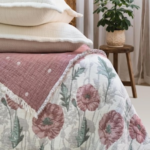 May include: A bed featuring a cream-coloured quilted coverlet with a floral pattern of pink poppies and sage green leaves, topped with a folded dusty rose muslin blanket with white polka dots and two cream-coloured ruffled muslin pillows.