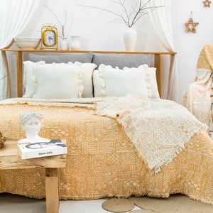 Könnte beinhalten: Eine gelbe und weiße gemusterte Tagesdecke mit Fransen auf einem Bett mit weißen Kissen. Die Decke ist zurückgeschlagen, um eine weiße und gelbe gemusterte Bettdecke zu zeigen. Eine weiße Büste einer Frau steht auf einem Holztisch neben einem Buch mit dem Titel "Steve Jobs".