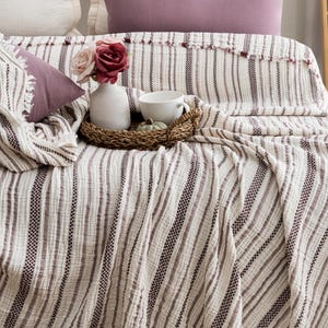 Pu&ograve; includere: Un letto con una coperta a righe color crema e cuscini abbinati. Un vassoio decorativo contiene una tazza bianca, una piccola zucca e un vaso con fiori. La coperta ha un bordo sfrangiato ed &egrave; drappeggiata sul letto.