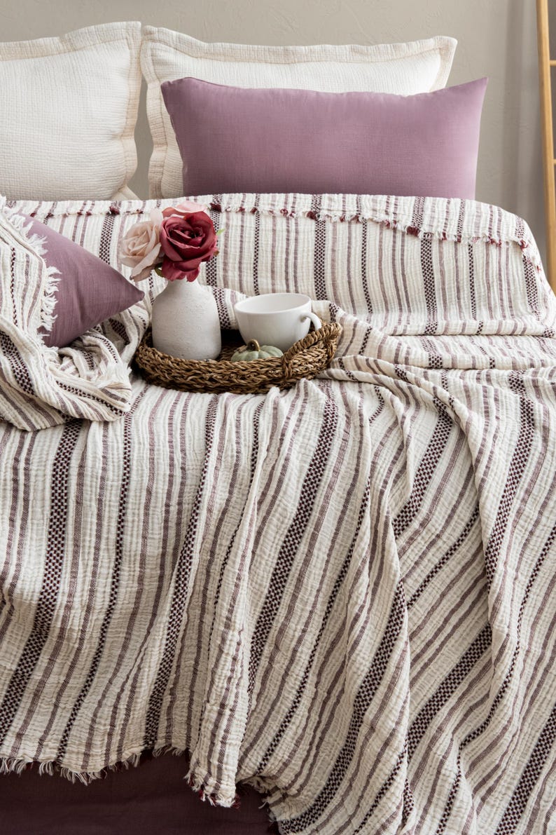 Pu&ograve; includere: Un letto con una coperta a righe color crema e cuscini abbinati. Un vassoio decorativo contiene una tazza bianca, una piccola zucca e un vaso con fiori. La coperta ha un bordo sfrangiato ed &egrave; drappeggiata sul letto.