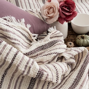 Pu&ograve; includere: Una coperta color crema con strisce bordeaux e frange &egrave; drappeggiata su una superficie. Sono presenti anche un cuscino malva, un vaso con rose rosa e bordeaux e una ciotola bianca, creando una scena accogliente e invitante.