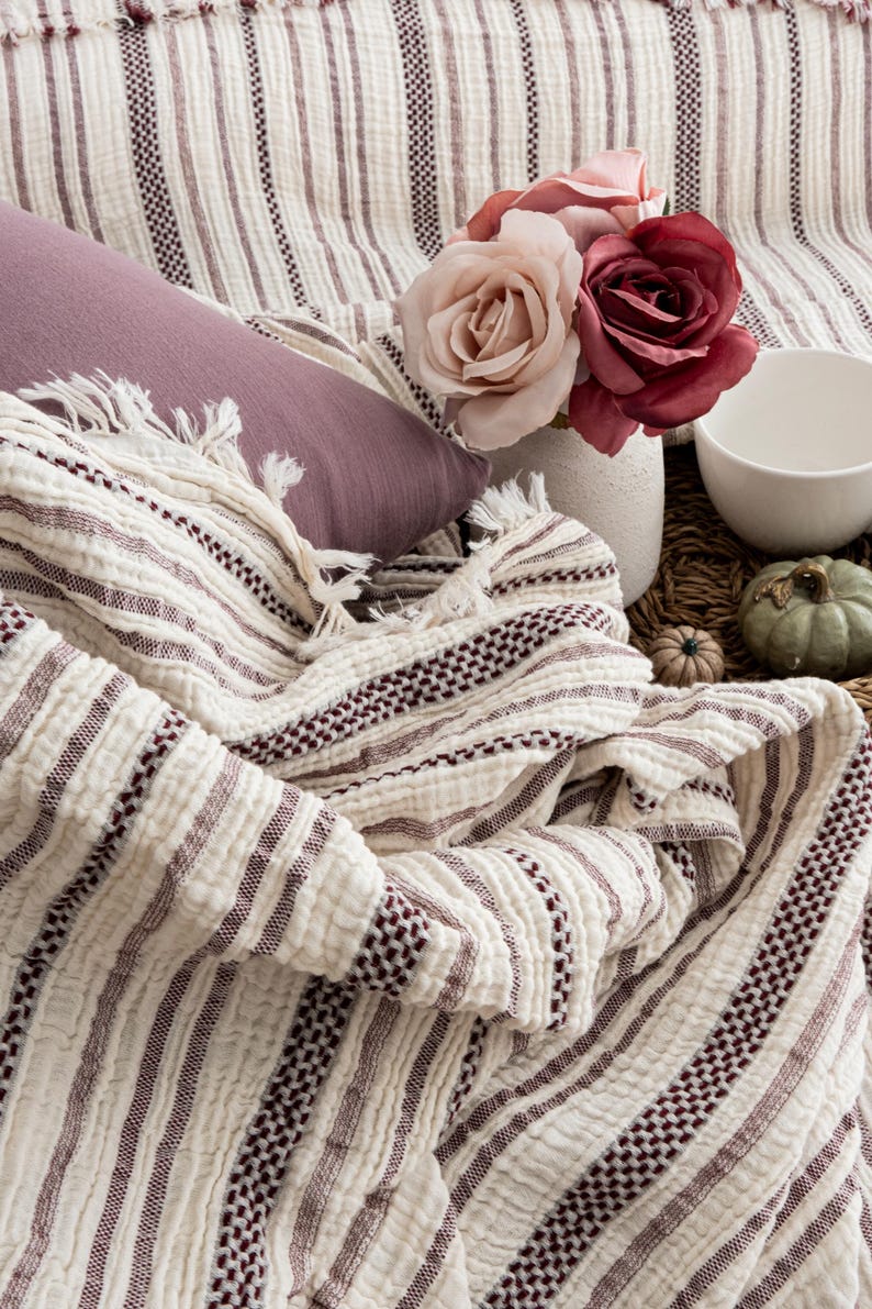 Pu&ograve; includere: Una coperta color crema con strisce bordeaux e frange &egrave; drappeggiata su una superficie. Sono presenti anche un cuscino malva, un vaso con rose rosa e bordeaux e una ciotola bianca, creando una scena accogliente e invitante.