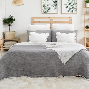 Könnte beinhalten: Eine graue Waffel-Decke mit ausgefransten Kanten, drapiert über ein Bett mit weißen Kissen und einer weißen Decke. Das Bett hat ein Kopfteil aus Holz und steht in einem Schlafzimmer mit einer weißen Backsteinwand und Pflanzen.