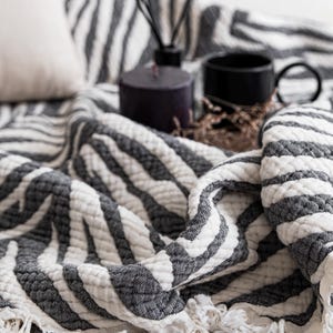 Op de afbeelding: Een getextureerde plaid met een contrastrijk zwart-wit gestreept patroon geïnspireerd op een zebra, afgewerkt met franjes. De stof heeft een gewatteerd, geplooid uiterlijk, wat zorgt voor een zachte, gelaagde uitstraling over het textieloppervlak.