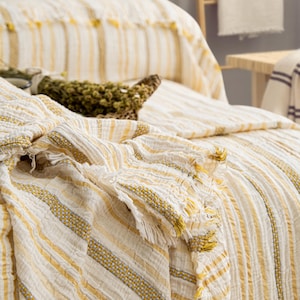 Pu&ograve; includere: Coperta a righe color crema con accenti gialli e frange. La coperta &egrave; drappeggiata su un letto, mostrando la sua trama strutturata e i dettagli decorativi. Un cestino intrecciato con erbe secche &egrave; appoggiato sul letto.