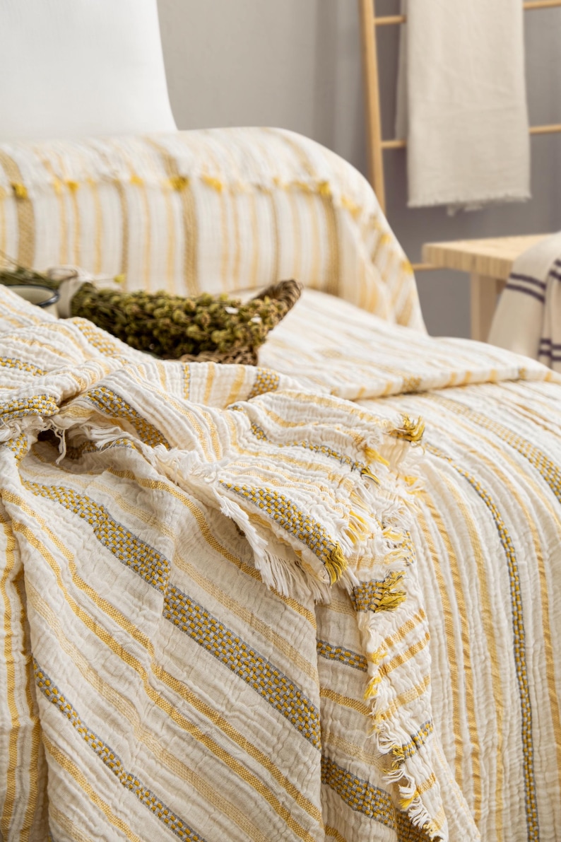 Pu&ograve; includere: Coperta a righe color crema con accenti gialli e frange. La coperta &egrave; drappeggiata su un letto, mostrando la sua trama strutturata e i dettagli decorativi. Un cestino intrecciato con erbe secche &egrave; appoggiato sul letto.