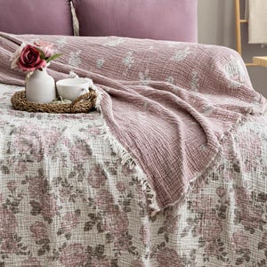 May include: A bed with a floral patterned blanket and matching pillows in shades of pink and white. A decorative tray with a vase of flowers and a cup sits on the bed. The blanket has a fringed edge.