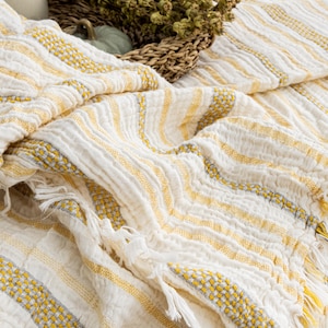 Pu&ograve; includere: Una coperta a righe bianche e gialle con frange. La coperta ha un aspetto strutturato con accenti gialli e grigi. Un cestino di vimini con erbe secche e una piccola zucca sono sullo sfondo.