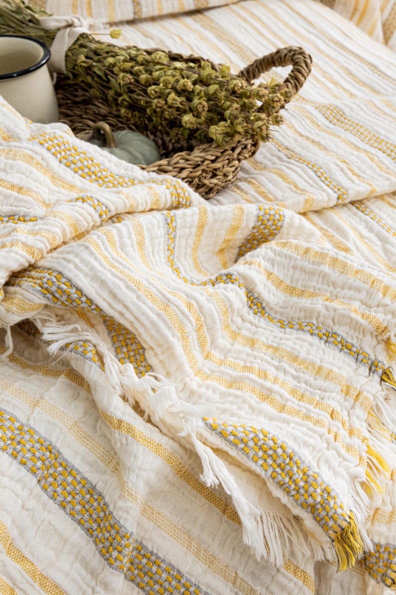 Pu&ograve; includere: Una coperta a righe bianche e gialle con frange. La coperta ha un aspetto strutturato con accenti gialli e grigi. Un cestino di vimini con erbe secche e una piccola zucca sono sullo sfondo.