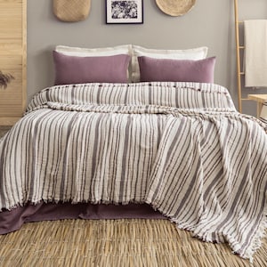 Pu&ograve; includere: Un letto con una coperta a righe e due cuscini malva. La coperta presenta strisce verticali nei toni del bianco e del marrone. Il letto &egrave; composto da un copriletto malva. La stanza ha una palette di colori neutri con texture naturali.
