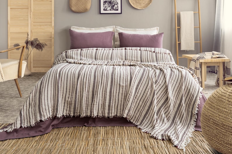 Pu&ograve; includere: Un letto con una coperta a righe e due cuscini malva. La coperta presenta strisce verticali nei toni del bianco e del marrone. Il letto &egrave; composto da un copriletto malva. La stanza ha una palette di colori neutri con texture naturali.