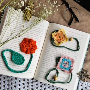 Könnte beinhalten: Drei gehäkelte Lesezeichen mit Blumenmotiven in Orange, Gelb und Pink mit blauen Akzenten. Jedes Lesezeichen hat einen grünen gehäkelten Stiel mit Blatt oder ein Holzperlendetail. Die Gegenstände liegen auf einem aufgeschlagenen Buch.
